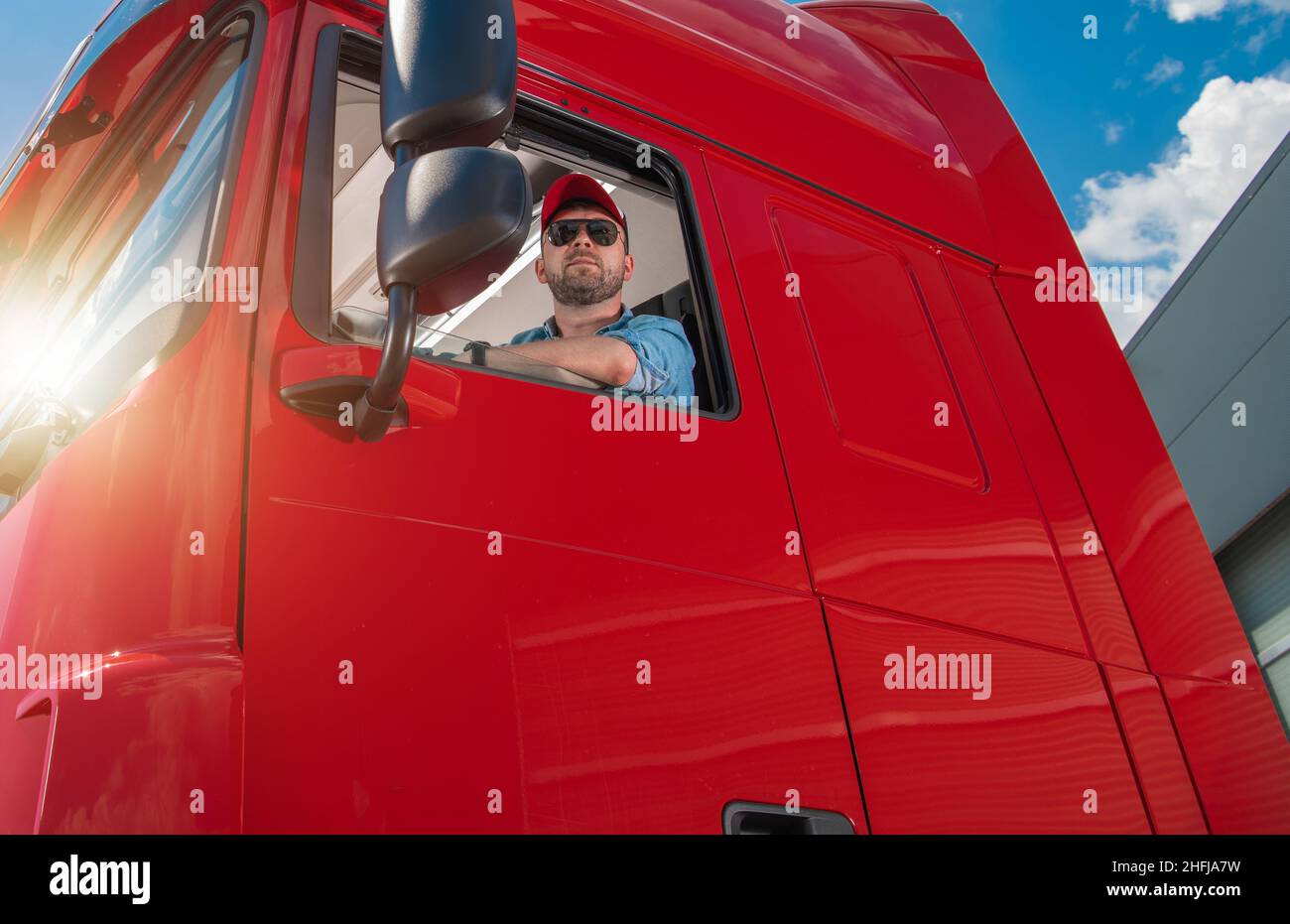 Professional Caucasian Trucker in His 30s Wearing Sunglasses Behind the Semi Tractor Truck Wheel. Commercial Transportation Industry. Stock Photo