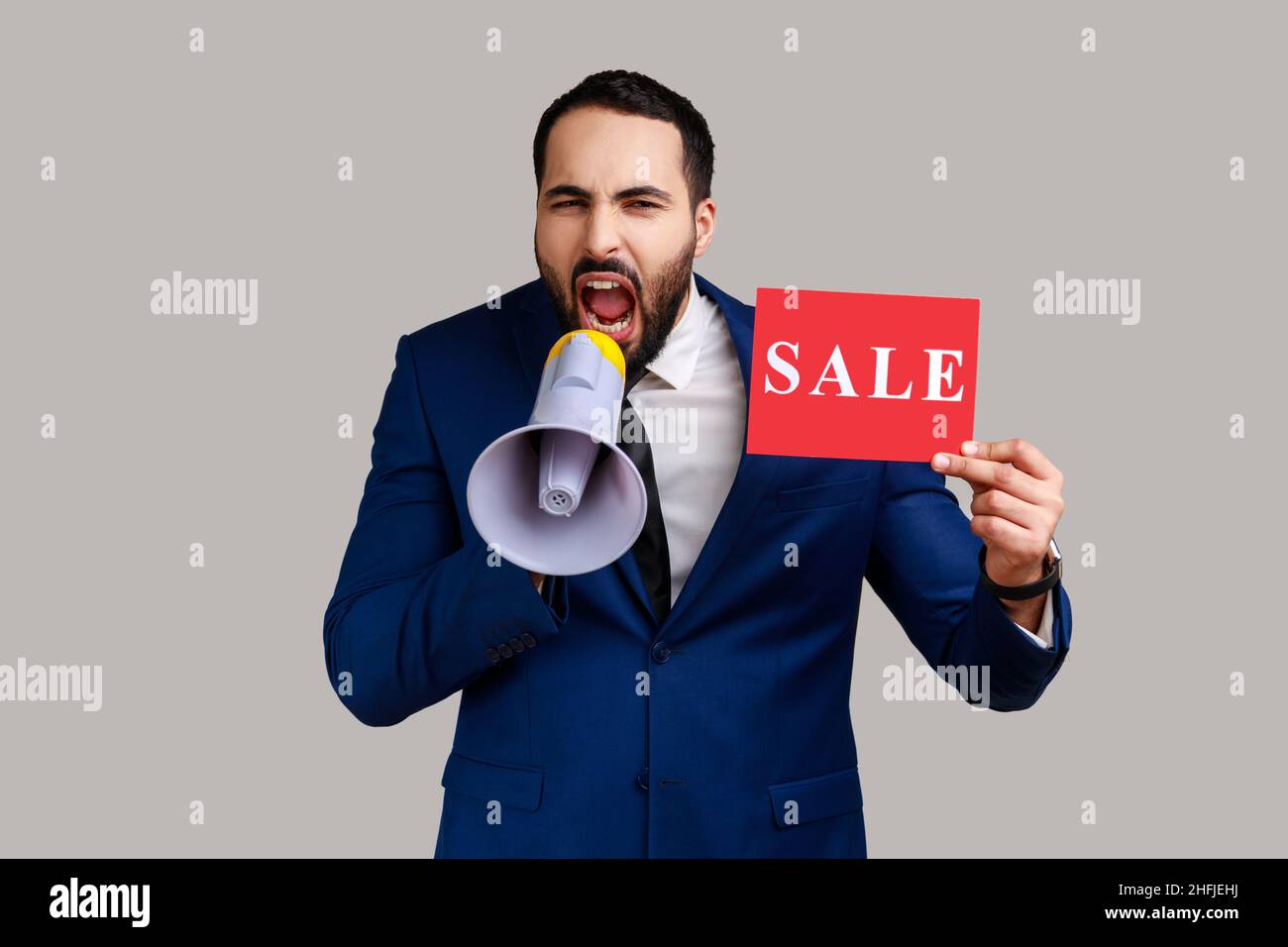 Portrait of bearded man paying attention announcing big sale yelling at loudspeaker, black Friday shopping, wearing official style suit. Indoor studio shot isolated on gray background. Stock Photo