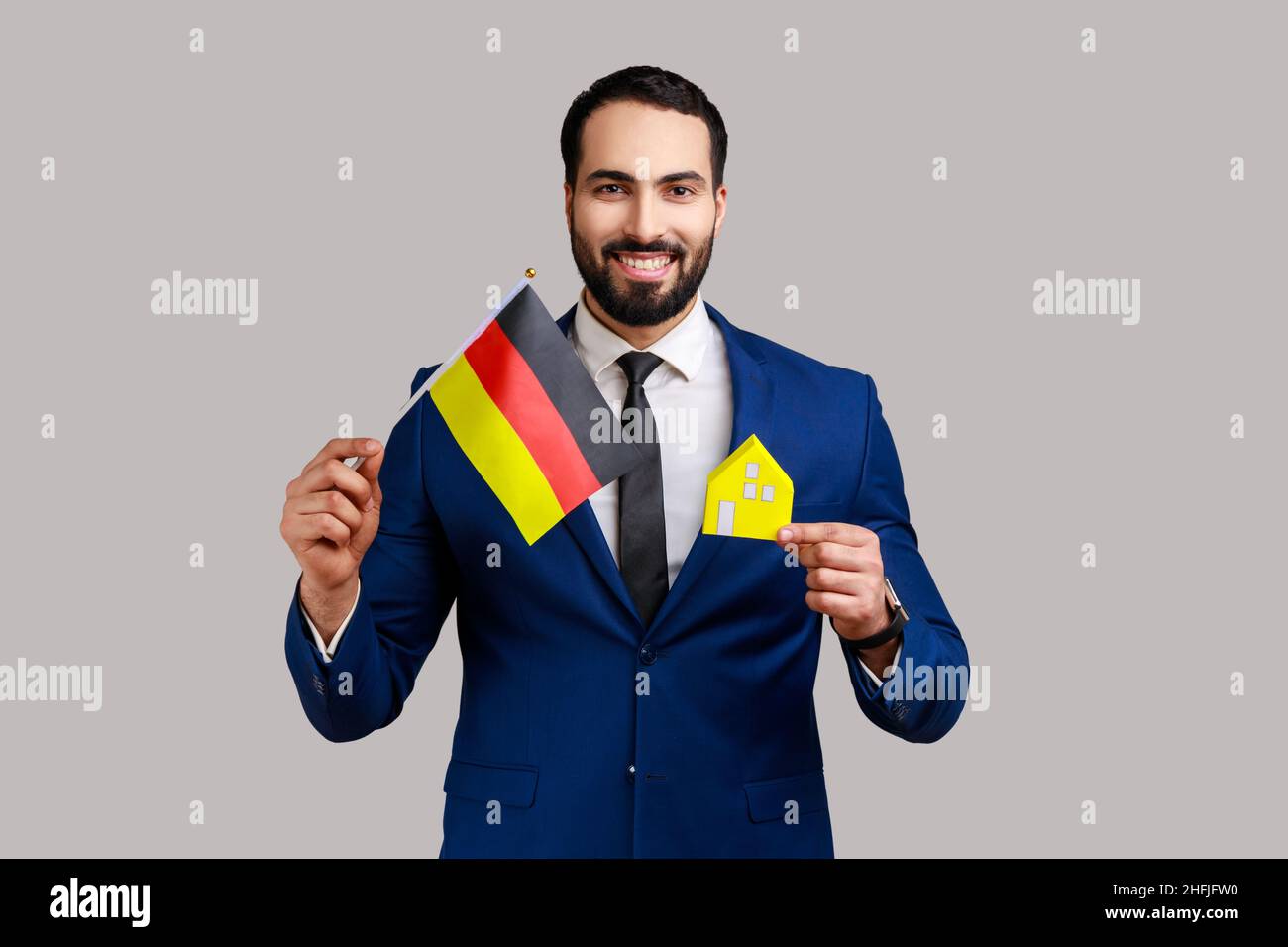 Satisfied man holding German flag and paper house, dreaming to buy his own house or flat in Germany, looking at camera, wearing official style suit. Indoor studio shot isolated on gray background. Stock Photo