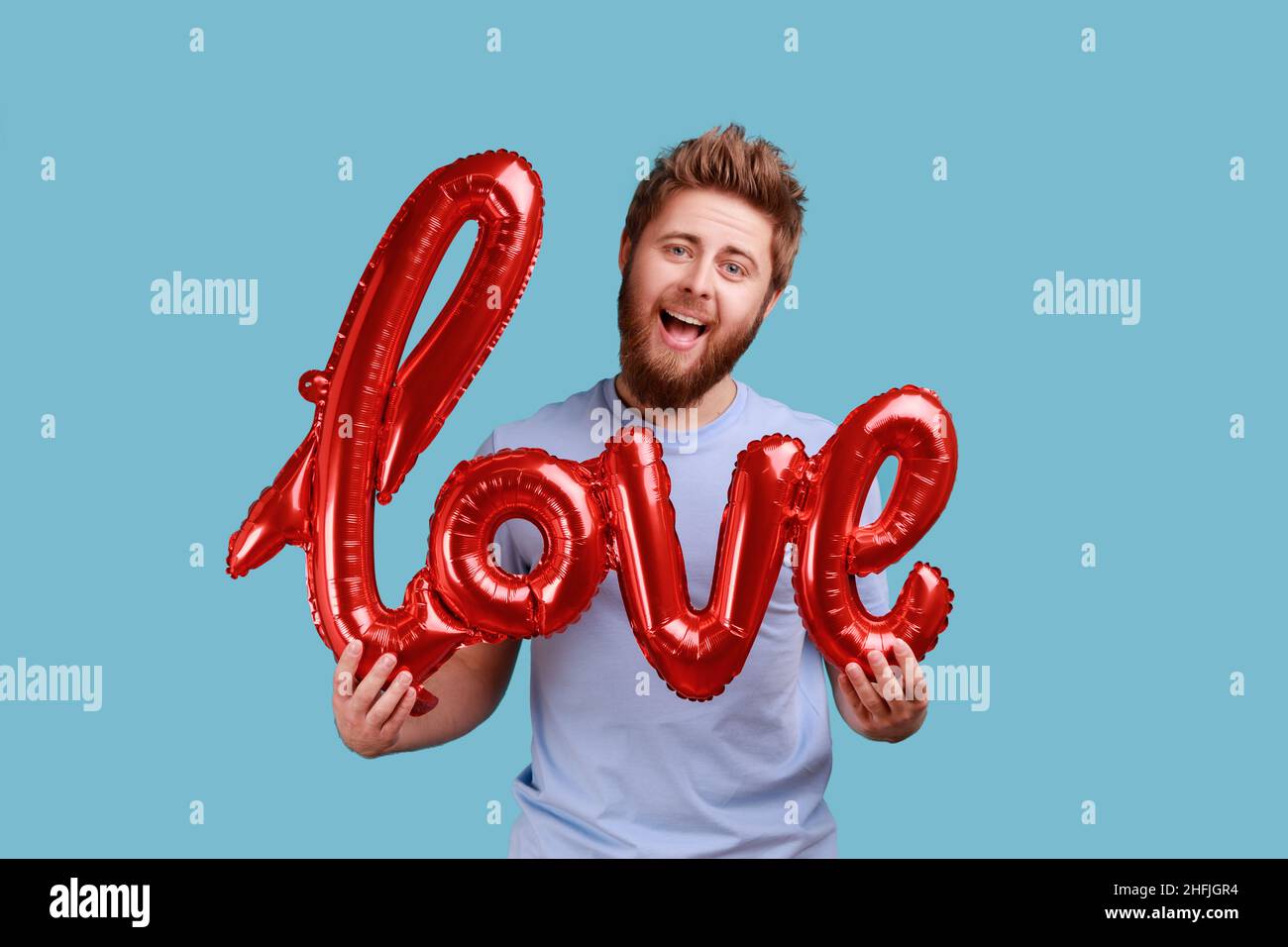 Portrait of romantic bearded man holding love word of foil balloons, expressing positive emotions and his feelings, being fall in love. Indoor studio shot isolated on blue background. Stock Photo