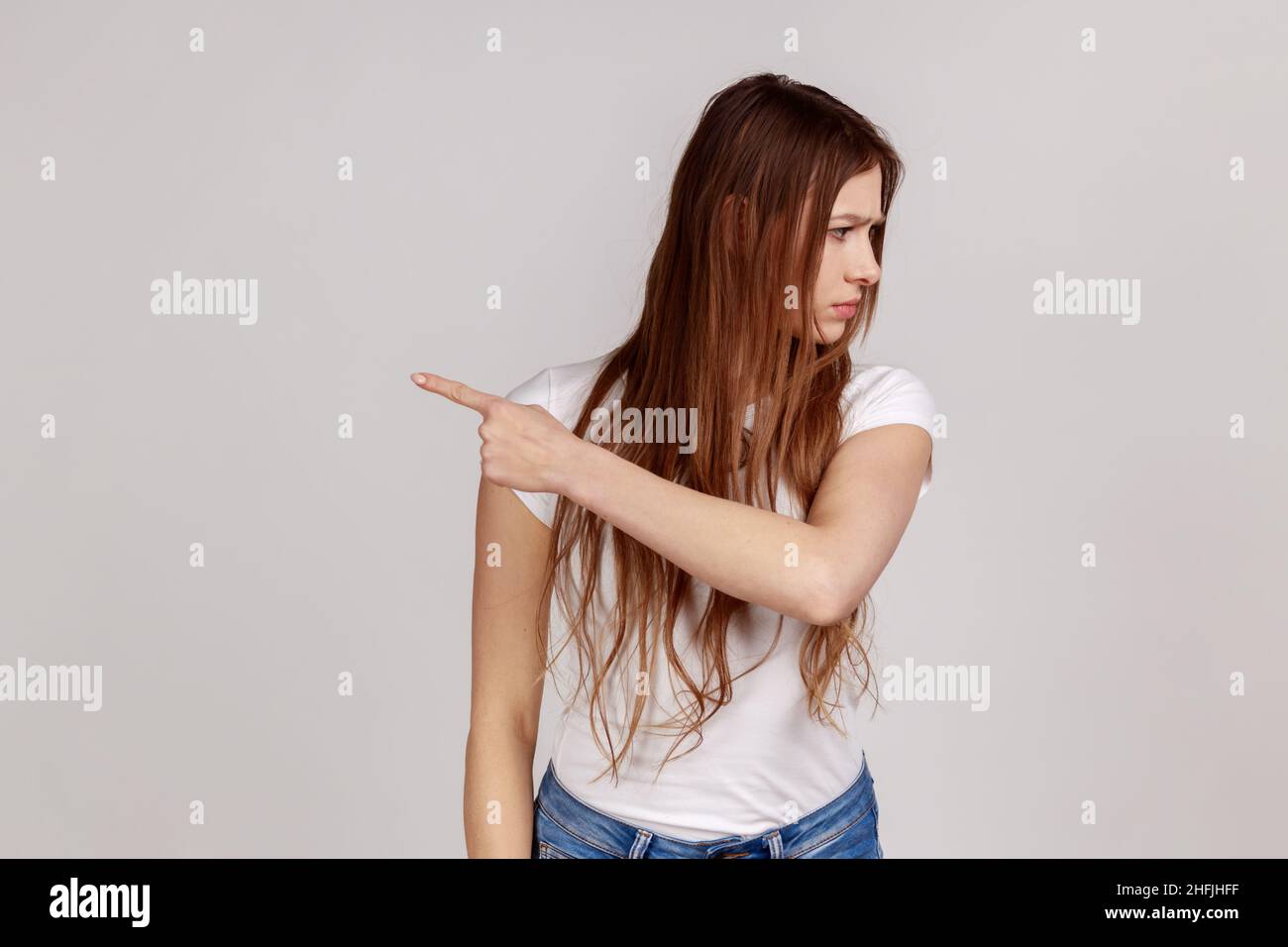 Portrait of resentful upset attractive woman turning away with disgust and ordering to leave, showing way out, feeling betrayed, wearing white T-shirt. Indoor studio shot isolated on gray background. Stock Photo