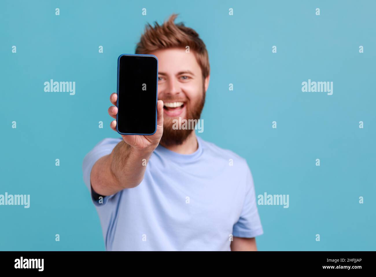 Portrait of bearded man holding out mobile phone and looking at camera with toothy smile and positive expression, being glad about technology. Indoor studio shot isolated on blue background. Stock Photo