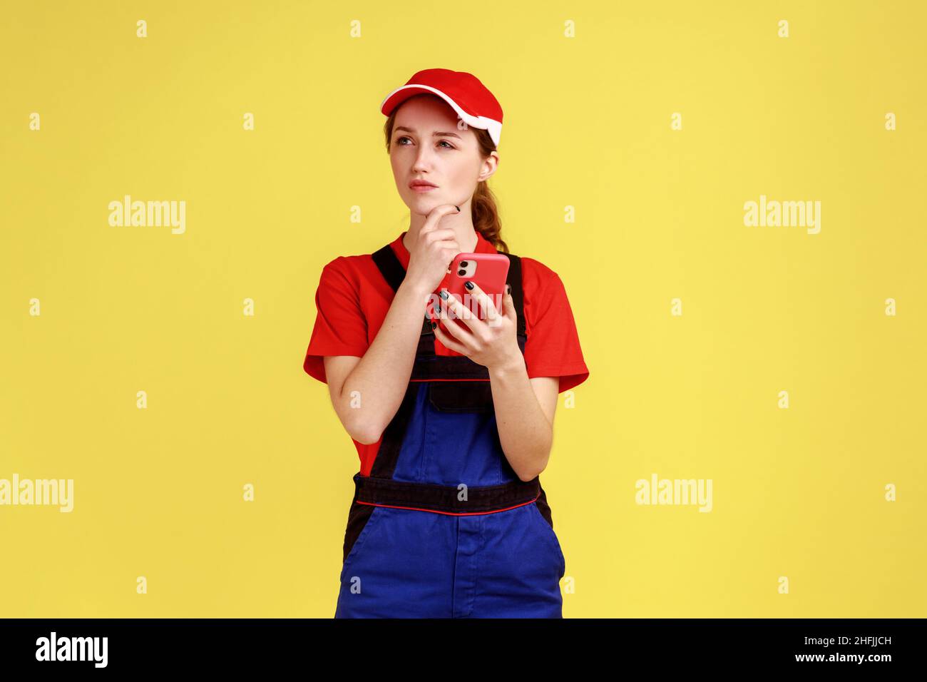 Portrait of thoughtful handy woman using mobile phone for online supporting, having pensive expression, holding chin and looking away Indoor studio shot isolated on yellow background. Stock Photo