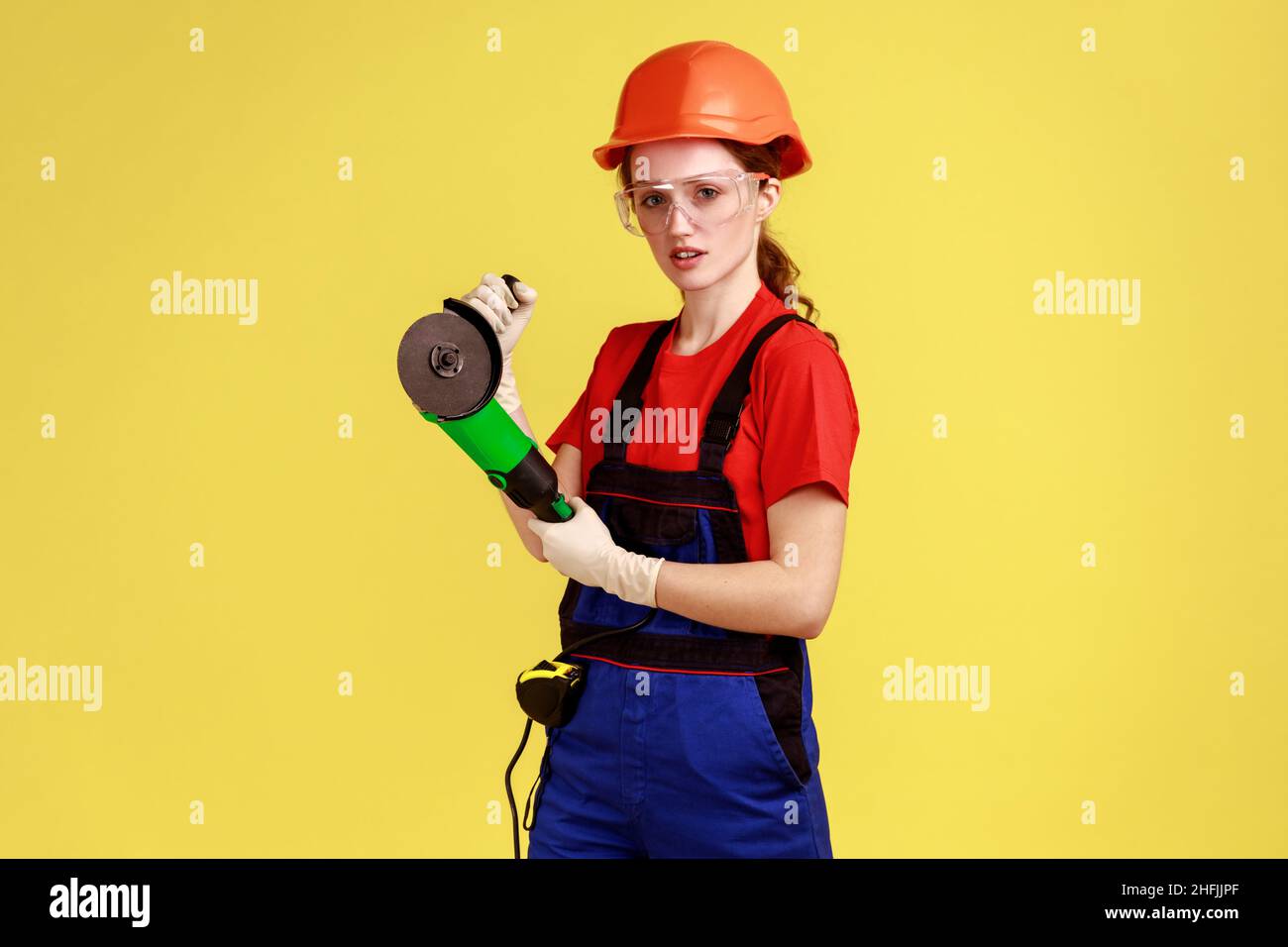 Portrait of serious carpenter woman working with angle grinder, having confident facial expressing, wearing overalls and protective helmet. Indoor studio shot isolated on yellow background. Stock Photo