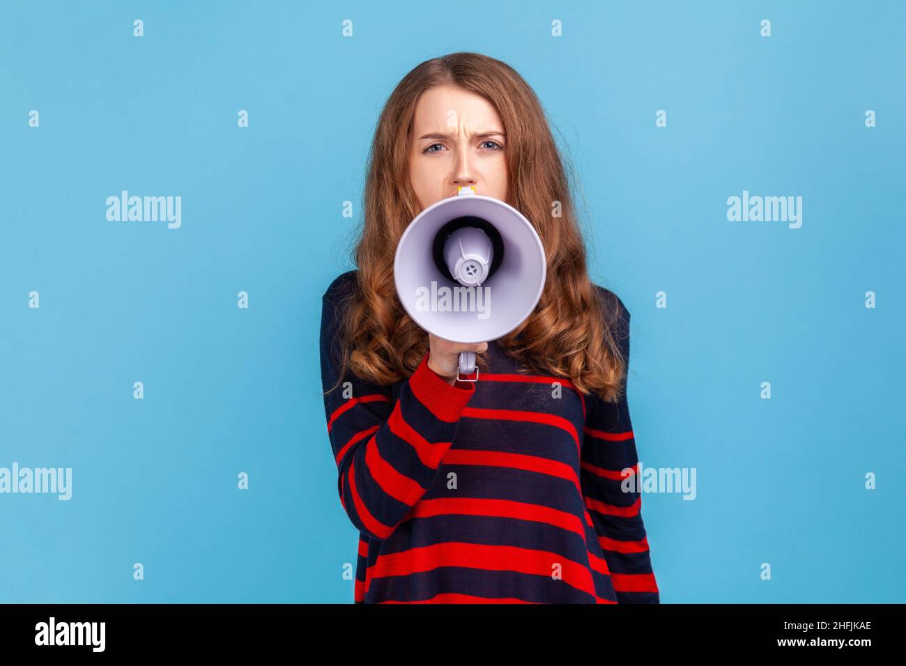 Portrait of woman wearing striped casual style sweater, taking in megaphone, looking at camera with serious expression, important announcement. Indoor studio shot isolated on blue background. Stock Photo