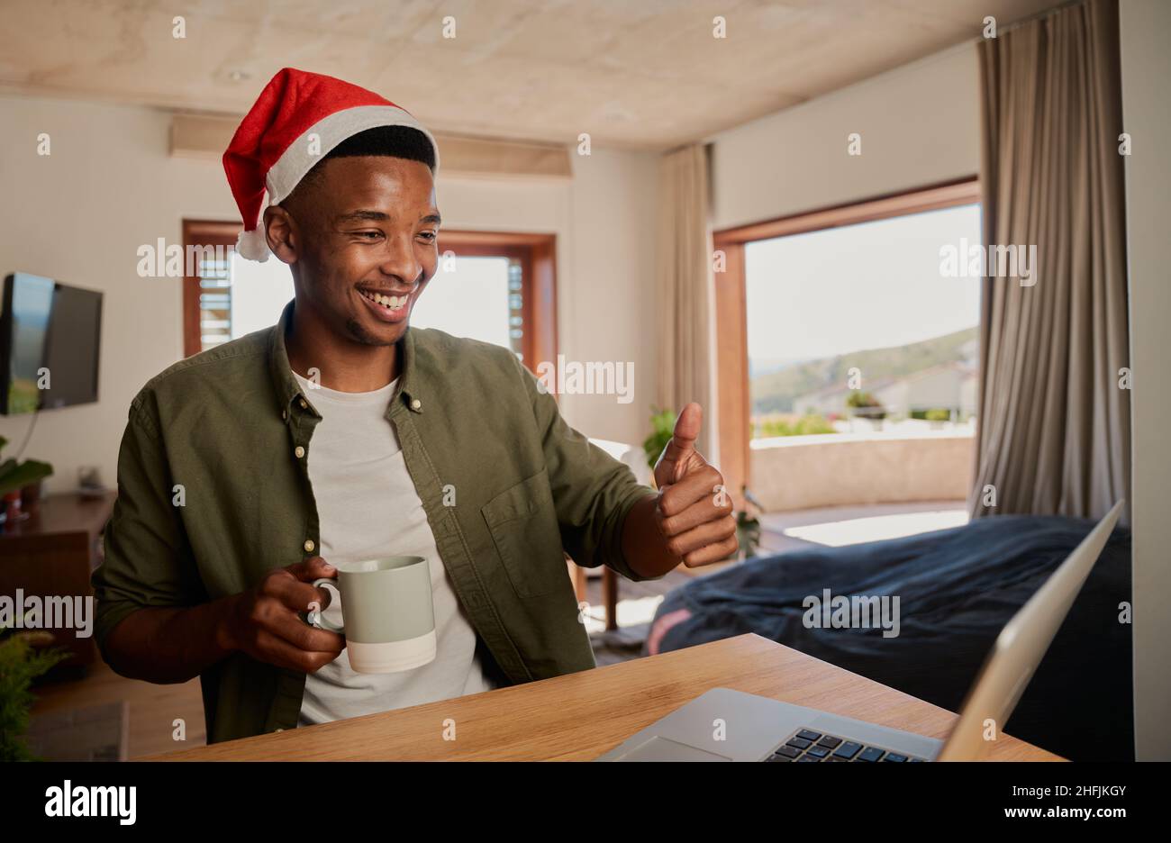 Young adult black male wearing Christmas hat, giving thumbs up while on online call. Sitting at kitchen counter using laptop. Stock Photo
