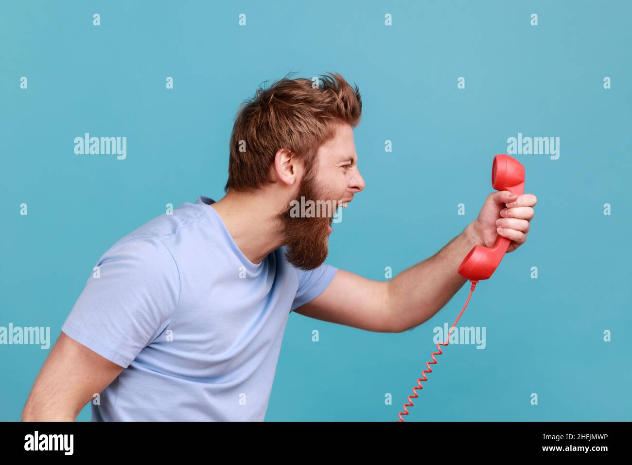 Side view portrait of handsome angry bearded man screaming and yelling talking retro landline phone, complaining on connection quality. Indoor studio shot isolated on blue background. Stock Photo