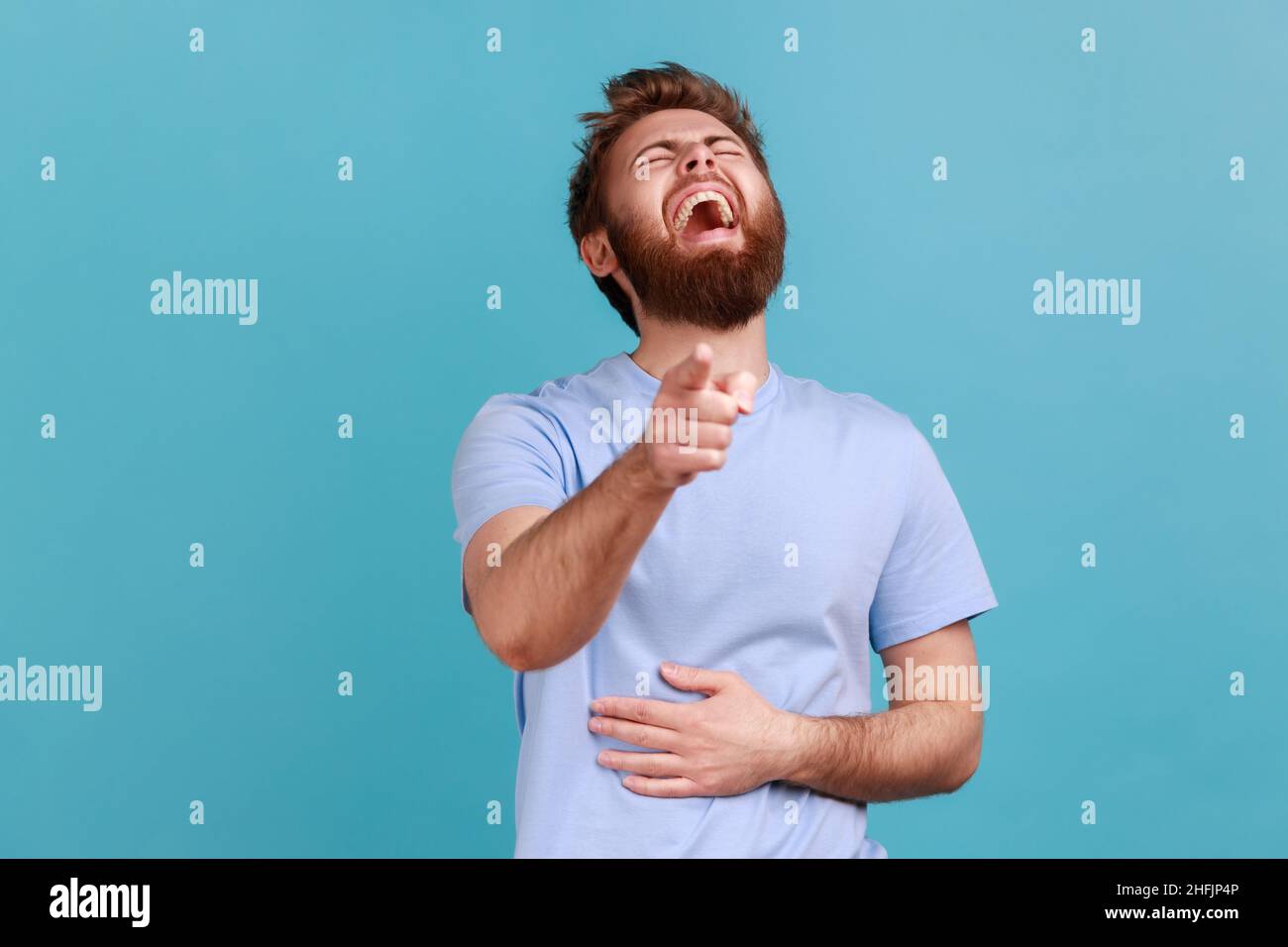 Hilarious laughter. Portrait of joyful happy bearded man laughing loudly and pointing to camera, mocking taunting you, holding belly. Indoor studio shot isolated on blue background. Stock Photo