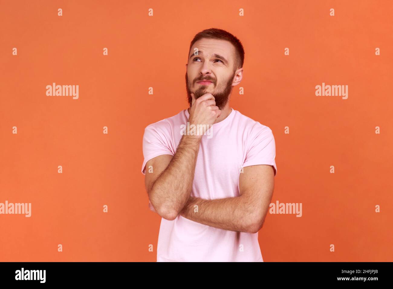 Portrait of thoughtful handsome bearded man holding his chin and pondering idea, confused not sure about solution, wearing pink T-shirt. Indoor studio shot isolated on orange background. Stock Photo