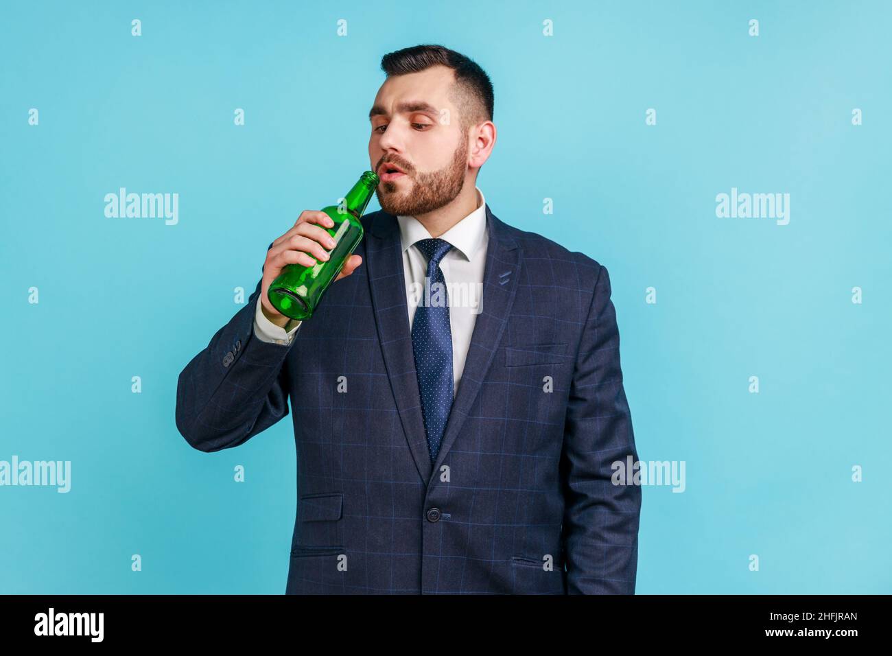 Bearded young adult businessman wearing official style suit drinking alcohol, holding beer bottle in hand, suffering from alcoholism. Indoor studio shot isolated on blue background. Stock Photo