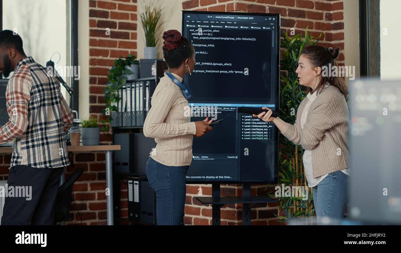 Programmer holding digital tablet analyzing code on wall screen tv while colleague is looking for errors pointing using remote control. Team of app developers collaborating on big data group project. Stock Photo