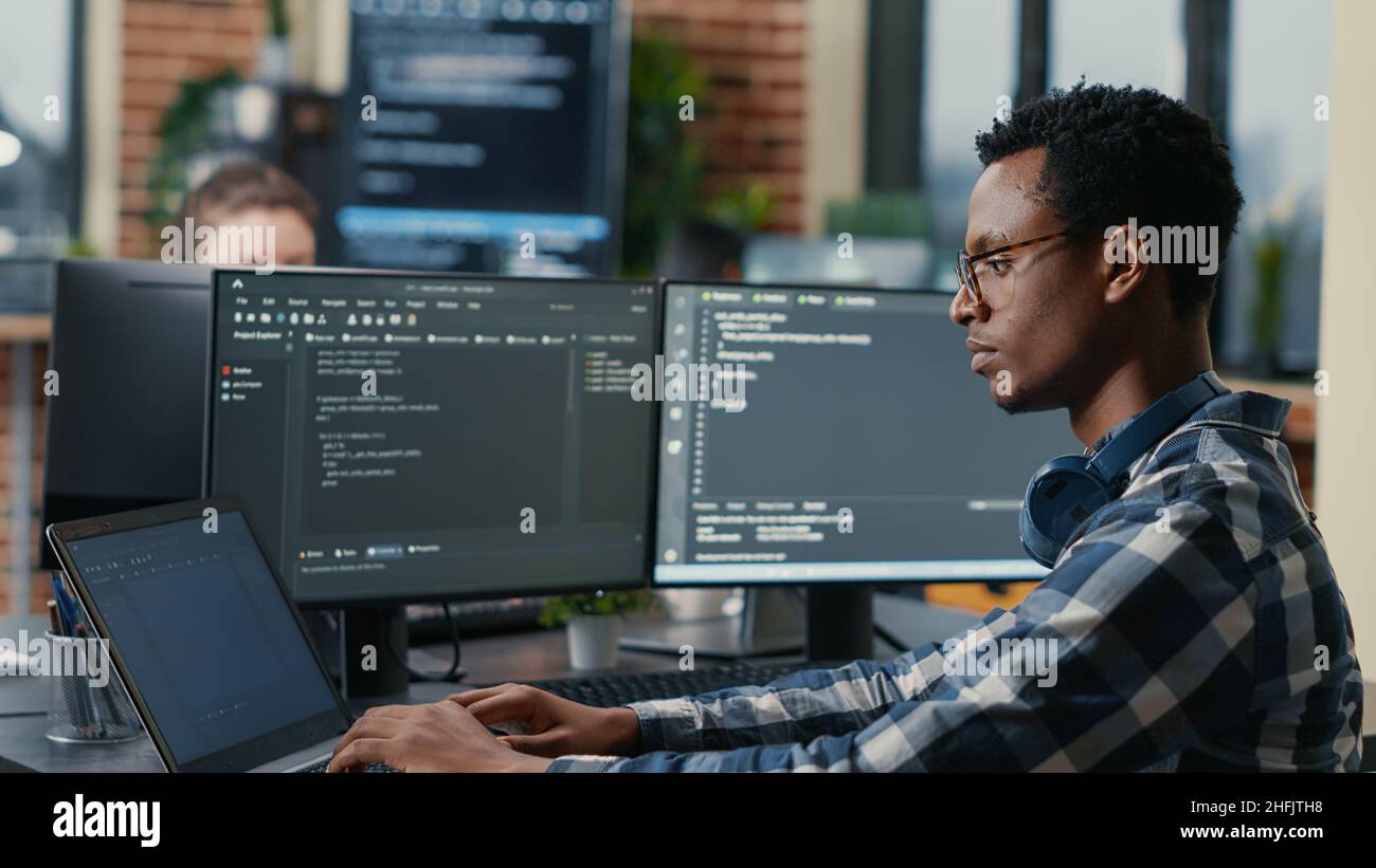 Sofware developer thinking while touching beard while typing on laptop sitting at desk with multiple screens parsing code. Focused database admin working with team coding in the background. Stock Photo