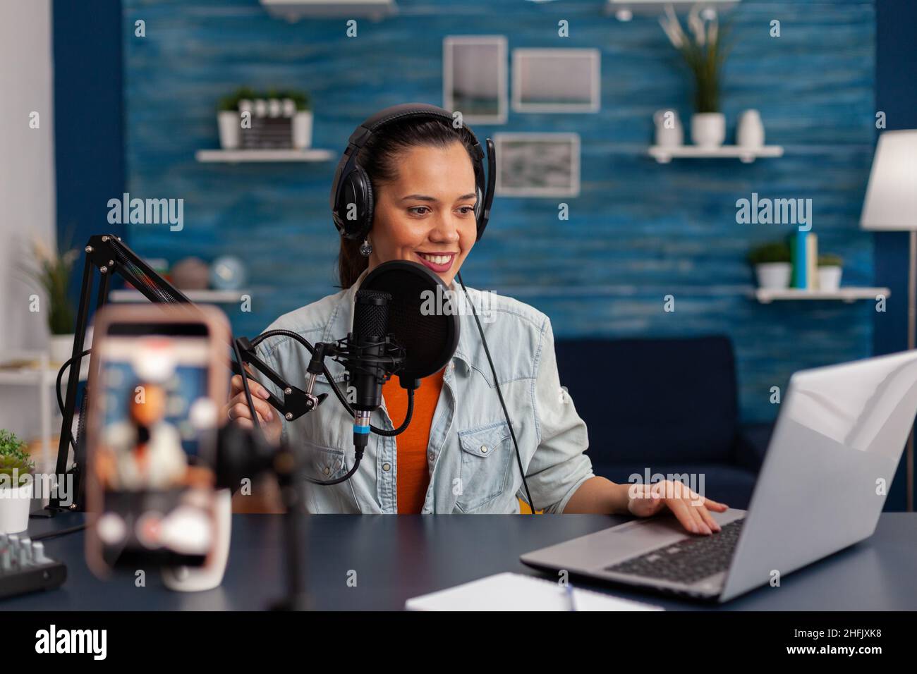 Woman vlogger with headphones looking at laptop and recording video. Influencer streaming online podcast on smartphone. Content creator in home studio using audio broadcasting equipment. Stock Photo