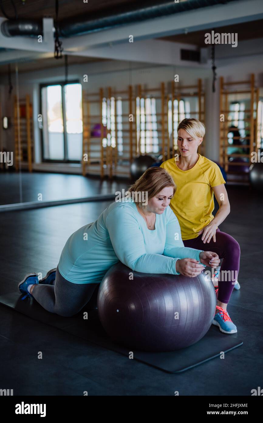 Overweight woman exercising with personal trainer on fintess ball in gym Stock Photo