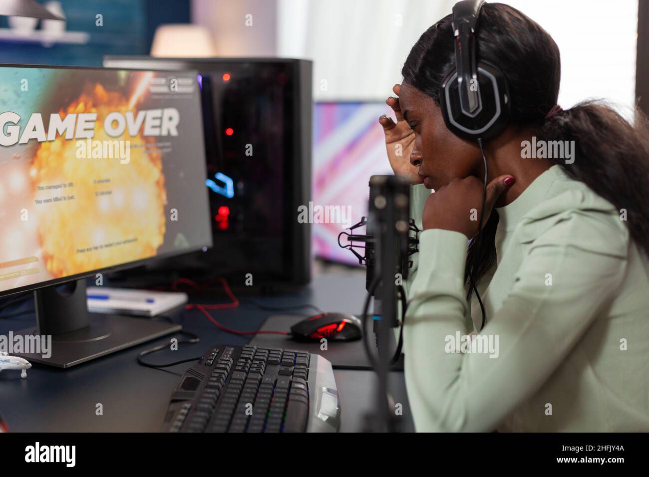 Sad upset woman gamer playing space shooter videogames losing online tournament while talking with players on streaming chat. Nervous streamer using RGB gaming computer. Game over on screen Stock Photo