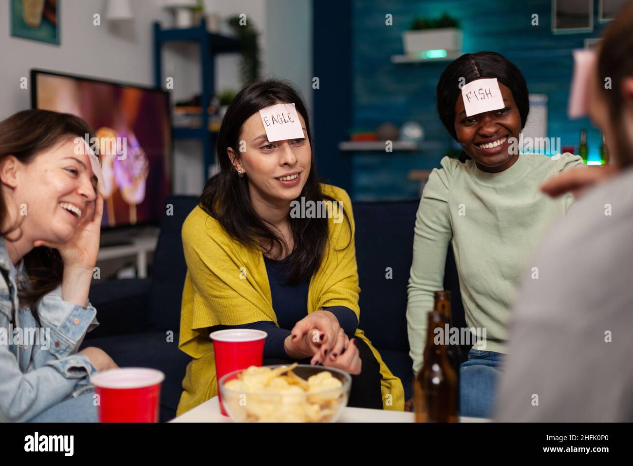 Multi-ethnic friends with stickey notes on forehead playing guess who game enjoying spending time together hanging out at home in living room. Group of people having fun during evening party Stock Photo