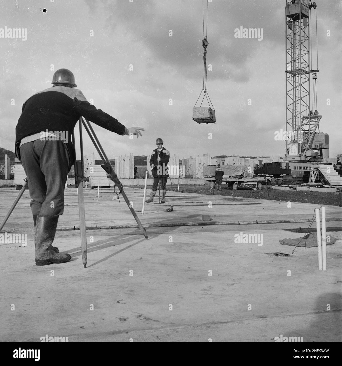 Craigshill, Livingston, West Lothian, Scotland, 01/10/1965. Two surveyors at work on a Jespersen construction compound, probably at the Craigshill development in Livingston, with a crane in the background unloading a pallet from a trailer. In 1963, John Laing and Son Ltd bought the rights to the Danish industrialised building system known as Jespersen (sometimes referred to as Jesperson). The company built factories in Scotland, Hampshire and Lancashire producing Jespersen prefabricated parts and precast concrete panels, allowing the building of housing to be rationalised, saving time and mone Stock Photo