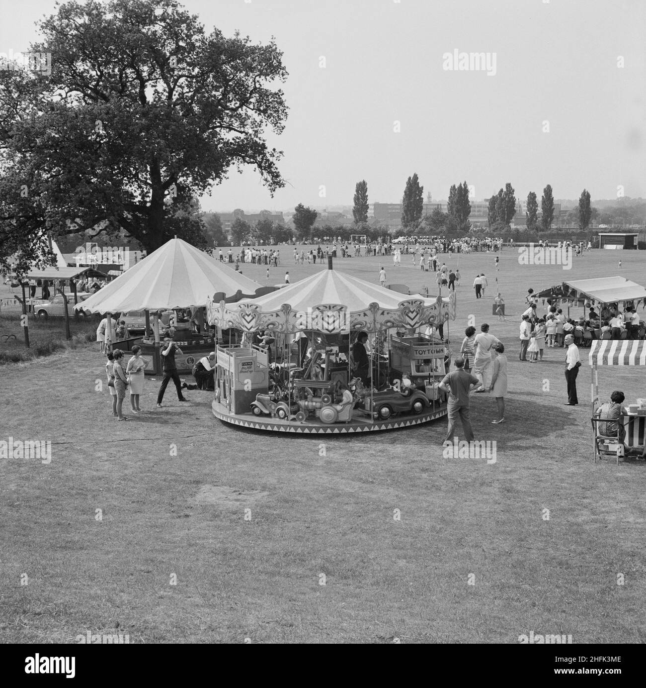Laing Sports Ground, Rowley Lane, Elstree, Barnet, London, 14/06/1969. A view of a fairground carousel and stalls, with crowds gathered on a sports pitch beyond, during a Gala Day held at the Laing Sports Ground at Elstree. A Gala Day was held by Laing at the Laing Sports Ground on 14th June 1969, as a replacement of the annual Sports Day. Sports events were held by the Sports Club, which included hockey, tennis, bowls, and football tournaments. A traditional English fete programme featured coconut shies, bingo, pony rides, catering and a beer tent, candy floss, and roundabouts. The day ended Stock Photo