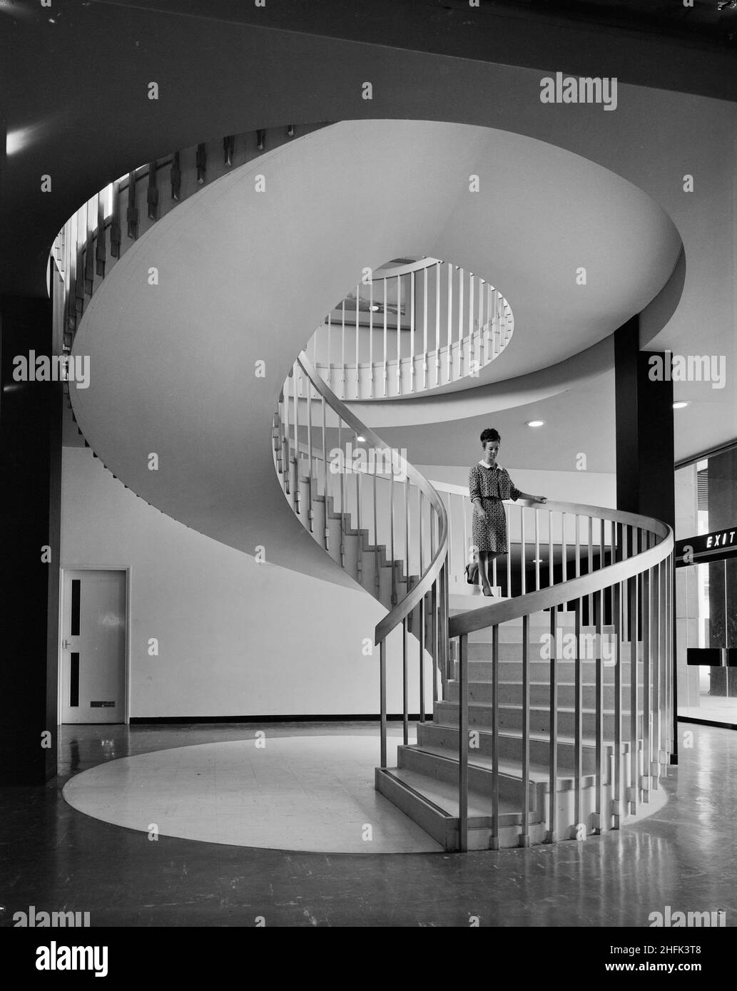 Paternoster Square, City of London, 23/06/1965. A woman walking down a winding staircase in a building at the Paternoster development. Work on the Paternoster development was carried out in a joint venture by John Laing Construction Limited, Trollope and Colls Limited, and George Wimpey and Company Limited. The scheme involved the redevelopment of a seven acre site on the north side of St Paul&#x2019;s Cathedral. The site had been almost entirely devastated during an incendiary raid in December 1940. The development consisted of a series of office blocks, a shopping precinct, an extensive piaz Stock Photo