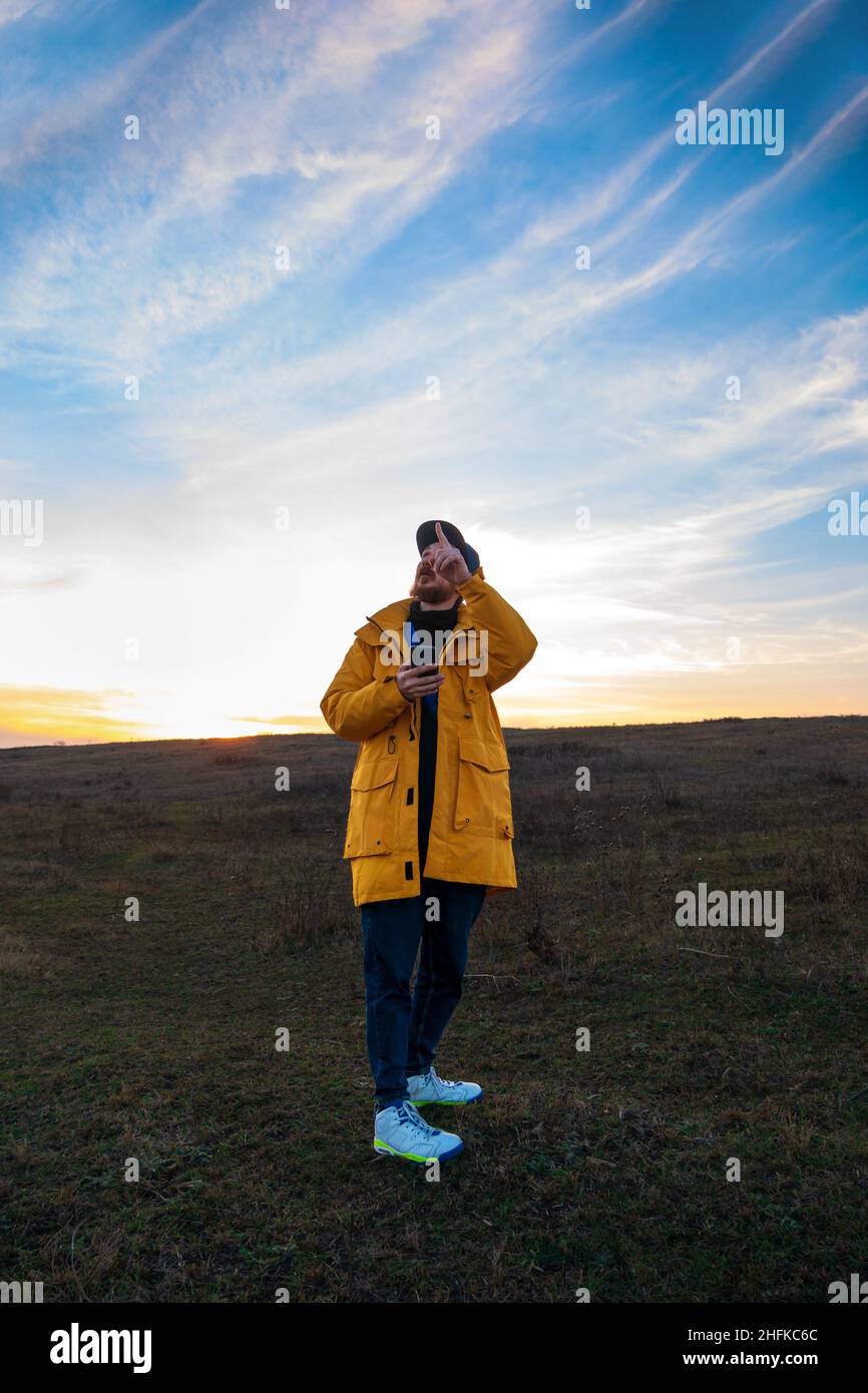 Traveler man in yellow coat with smartphone walking on a hill sunset sky side view. Authentic male tourist with gadget Stock Photo