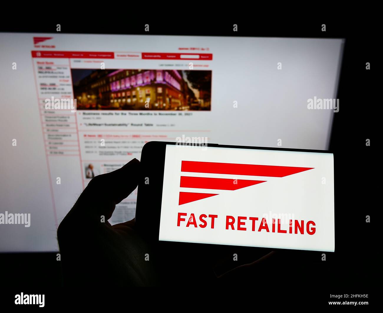 Person holding mobile phone with logo of Japanese retail company K.K. Fast Retailing on screen in front of web page. Focus on phone display. Stock Photo