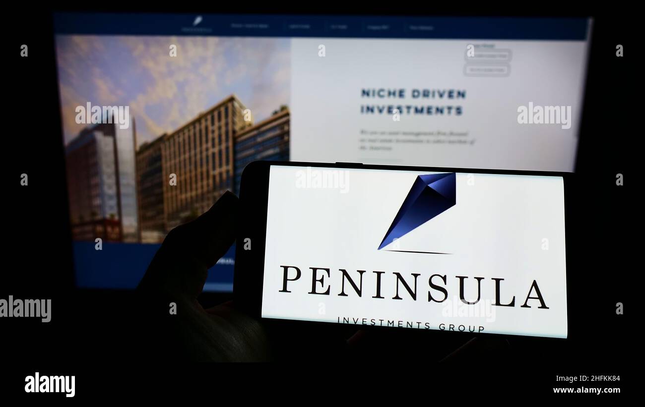 Person holding smartphone with logo of company Peninsula Investments Group Capital LLC on screen in front of website. Focus on phone display. Stock Photo