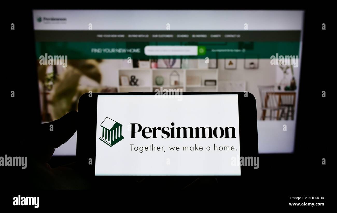 Person holding smartphone with logo of British housebuilding company Persimmon plc on screen in front of website. Focus on phone display. Stock Photo