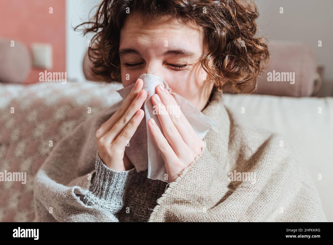 Sick upset young woman sitting on sofa covered with warm plaid, freezing, grimacing, runny nose, fever, sneezing into tissue, sick girl with flu sympt Stock Photo