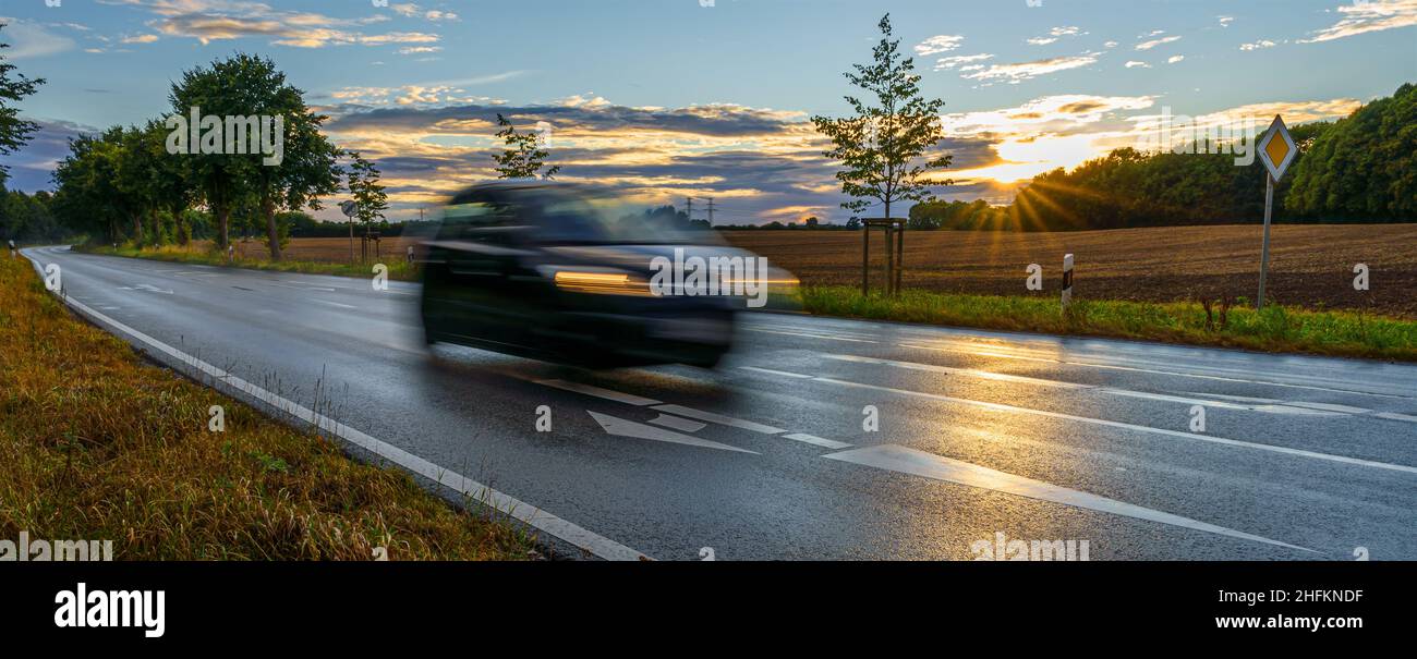Ein PKW fährt mit hoher Geschwindigkeit auf herbstlich nasser Straße; die Abendsonne scheint auf abgeerntete Felder Stock Photo