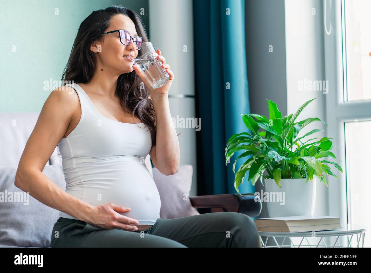 Pregnant woman with glasses sit on sofa caressing belly drinking glass of water Stock Photo