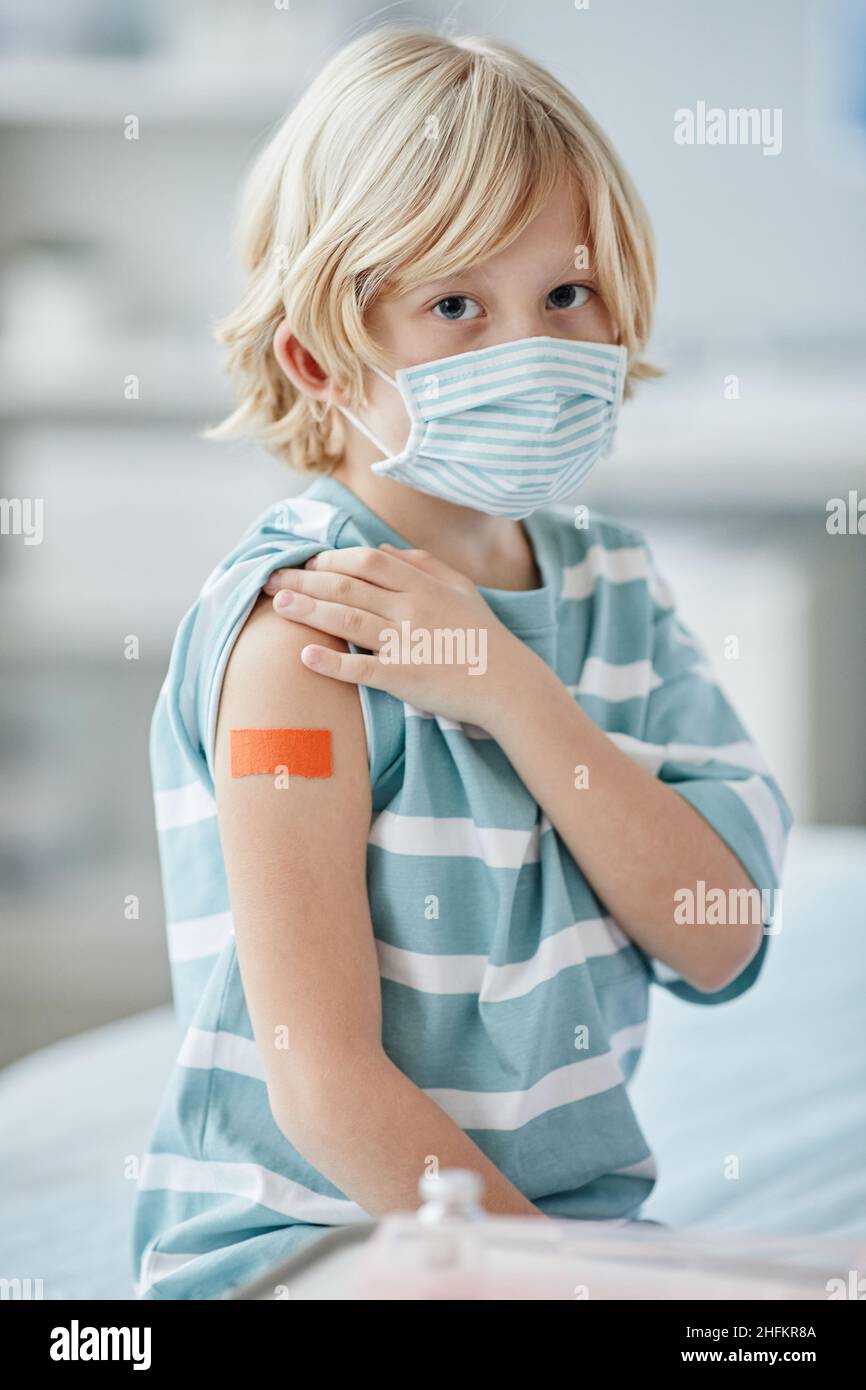 Vertical portrait of cute little boy showing shoulder patch after getting vaccinated against covid 19 in clinic Stock Photo