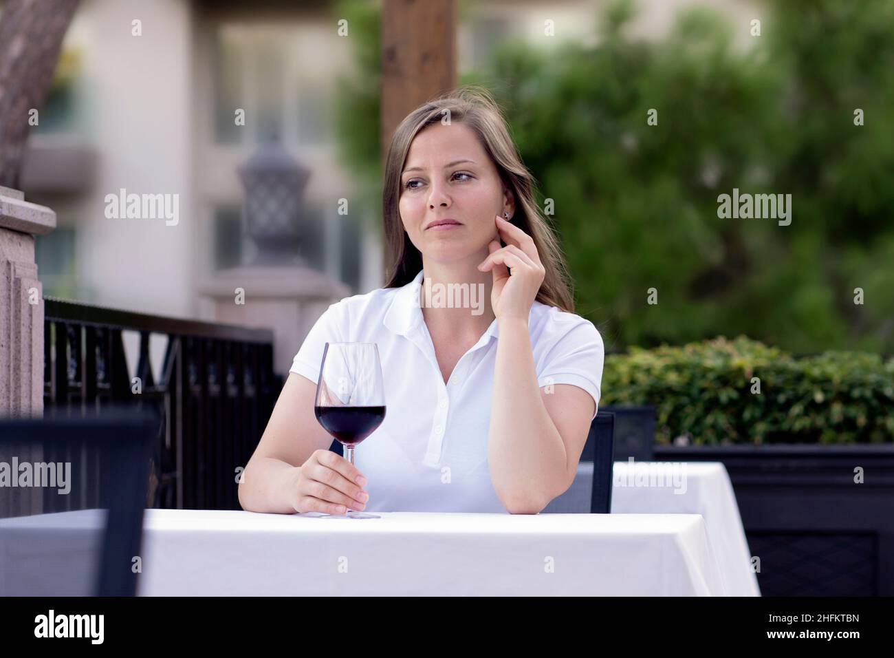 a woman is sitting alone at a table in a summer cafe or restaurant, holding a glass of red wine in her hands. expectation or a dreamy romantic mood. Stock Photo
