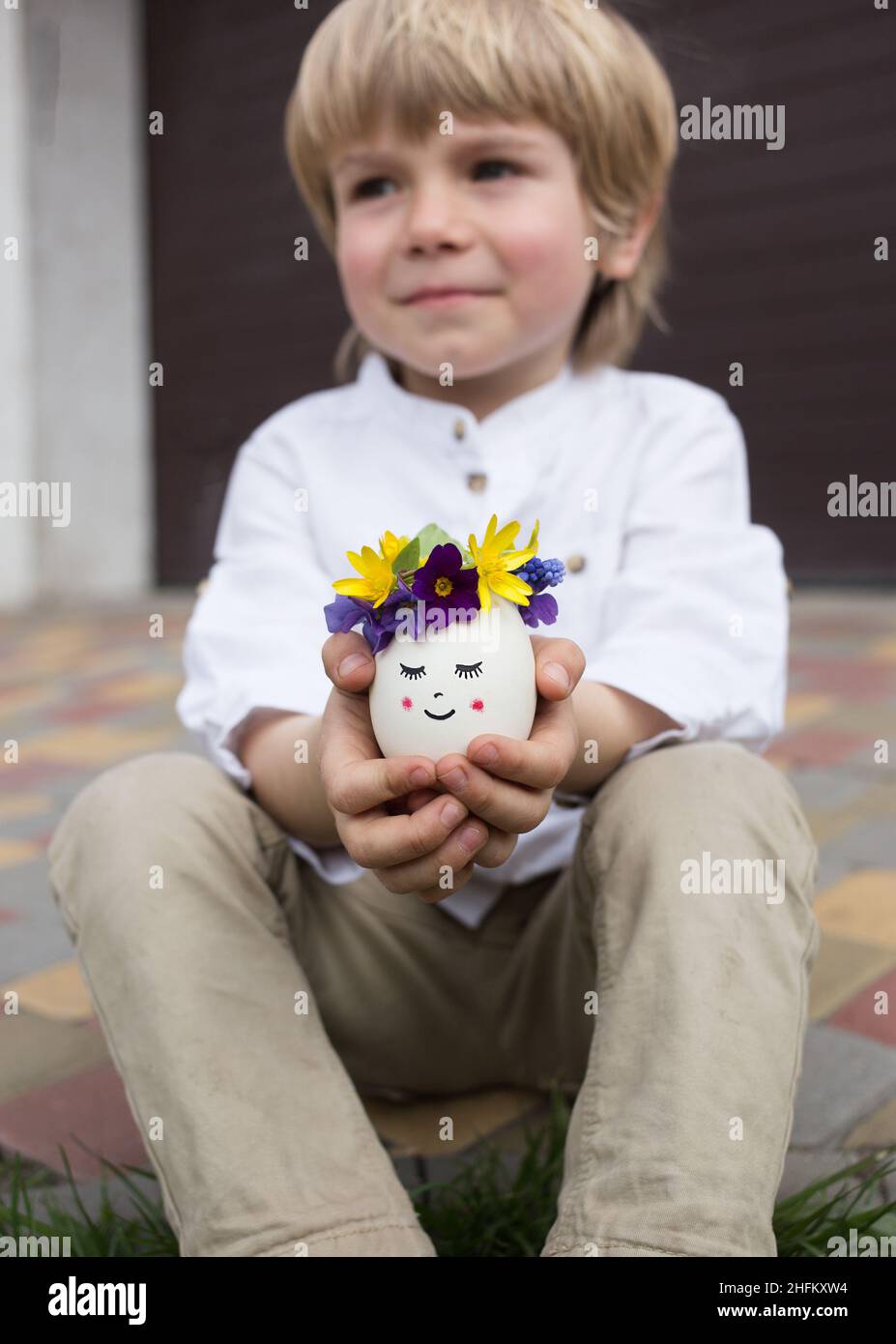 child hold easter egg in hands. egg is decorated with a wreath of fresh flowers and a cute face painted on it. Positive thinking, creative ideas. happ Stock Photo