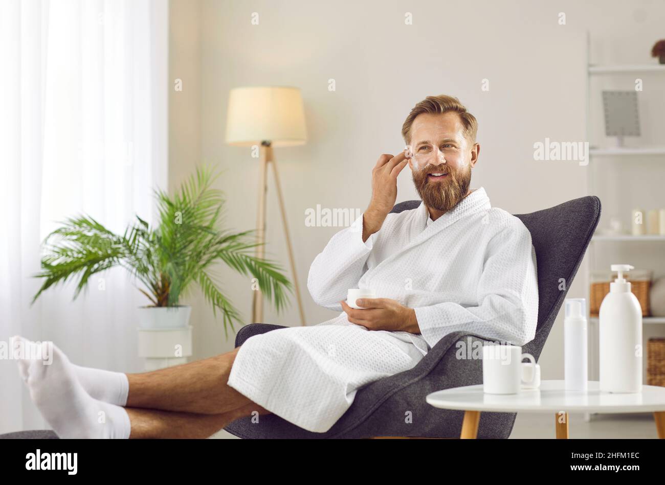 Funny bearded man puts cosmetic mask on his face while doing spa treatments at home. Stock Photo