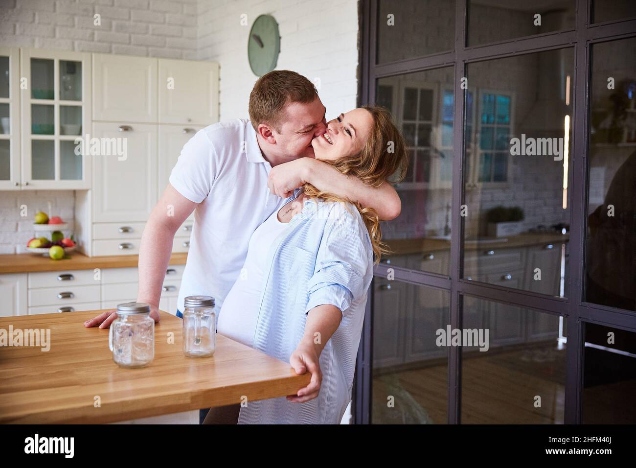 young pregnant woman and her husband hug and laugh merrily in the large kitchen of their house. Copy space, lifestyle. Stock Photo