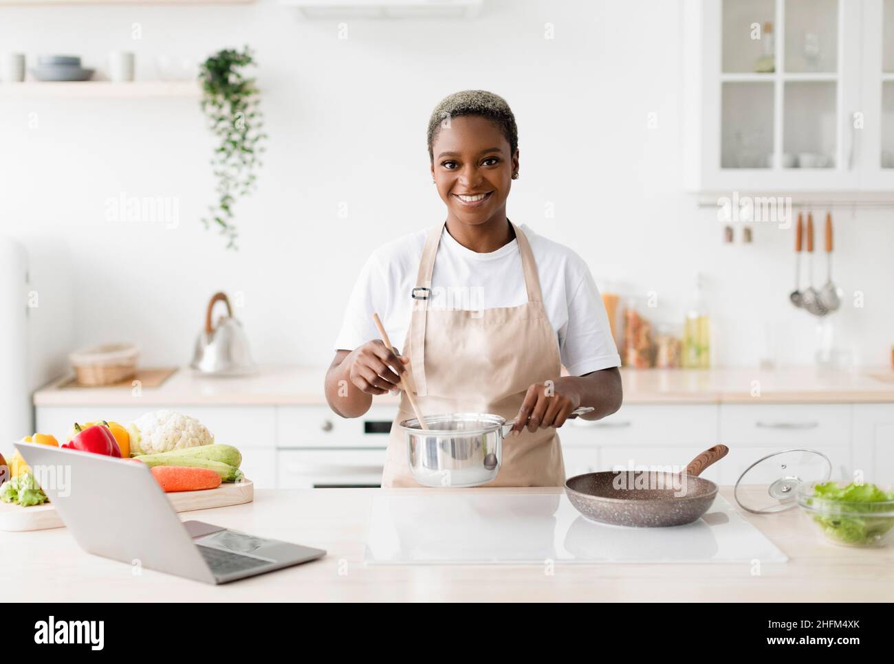 Happy young cute black lady in apron prepares lunch on table with pc in minimalist kitchen interior, free space Stock Photo