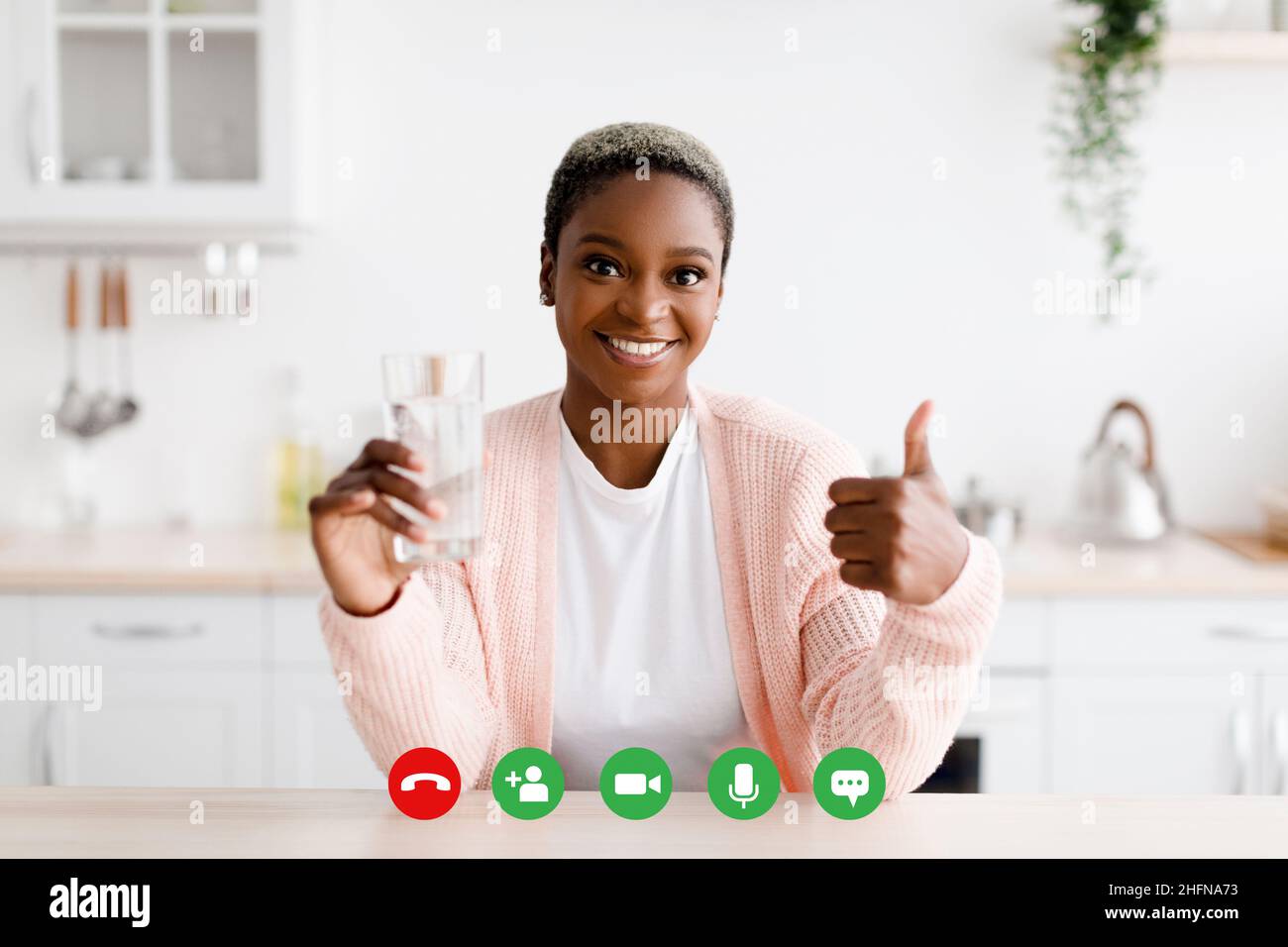 Happy millennial black female holds glass of clean water and shows thumb up on minimalist kitchen interior Stock Photo