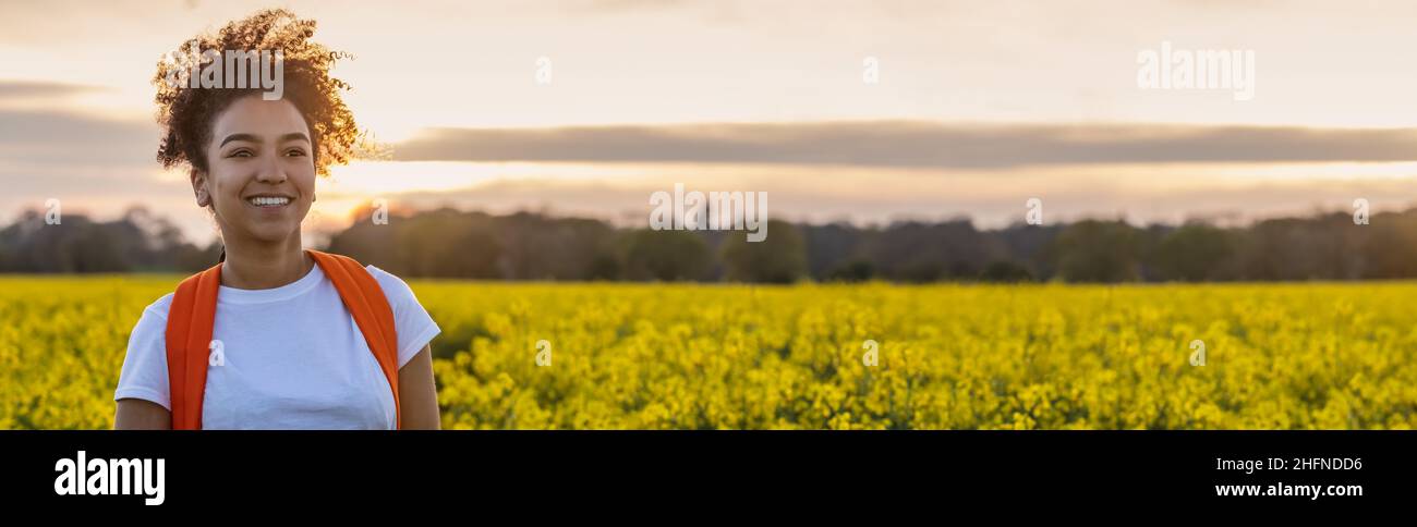 Panorama header beautiful mixed race African American girl teenager female young woman smiling and happy hiking with back pack in a field of yellow fl Stock Photo