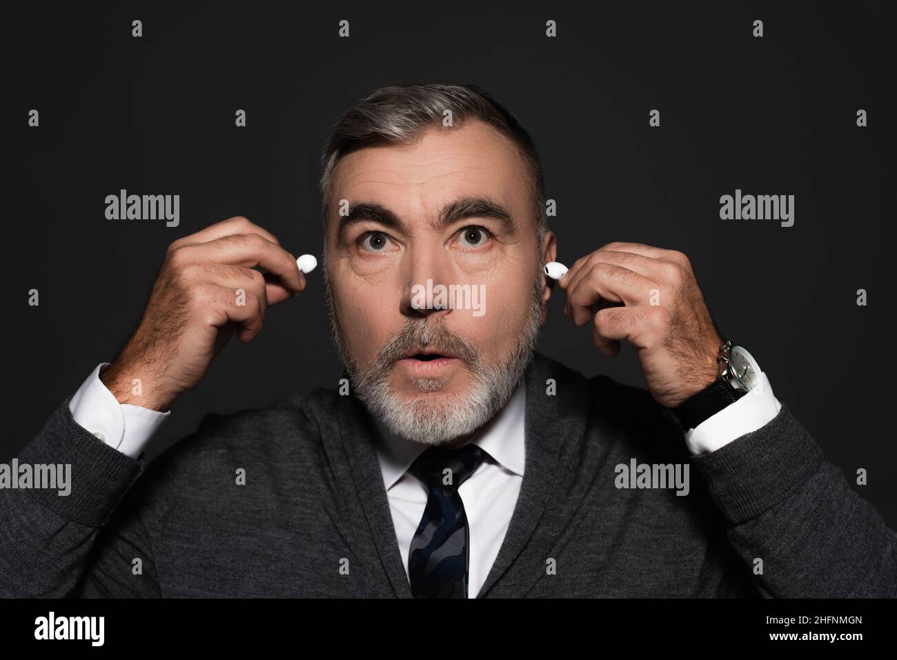 surprised senior man inserting earphones isolated on dark grey Stock Photo