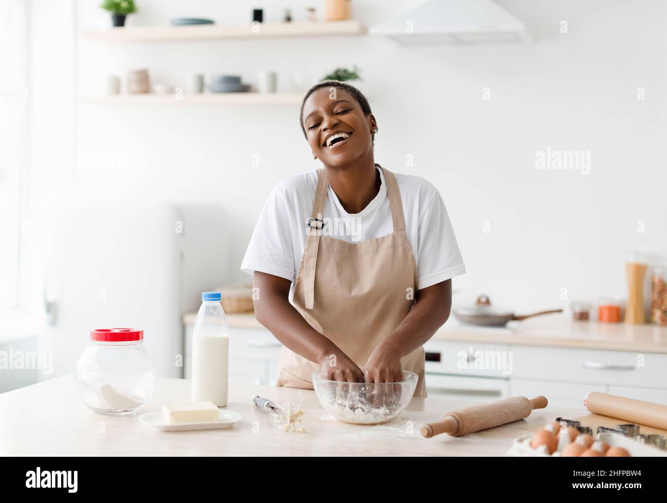 Happy glad laughing young attractive black lady in apron prepares dough for baking in modern kitchen interior Stock Photo