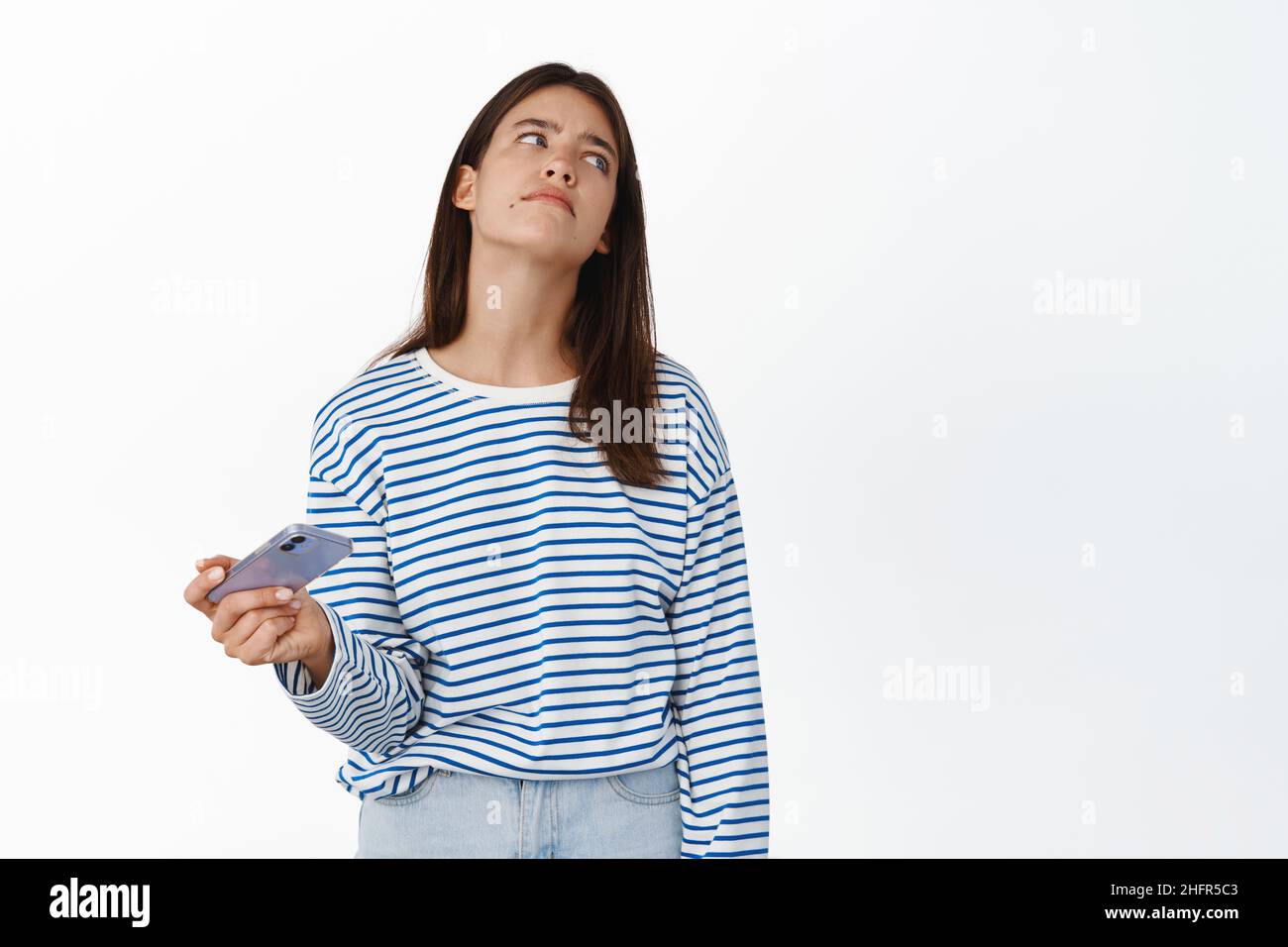 Sad and disappointed girl holding smartphone, losing in video game, sighing upset and looking away, standing uneasy against white background Stock Photo
