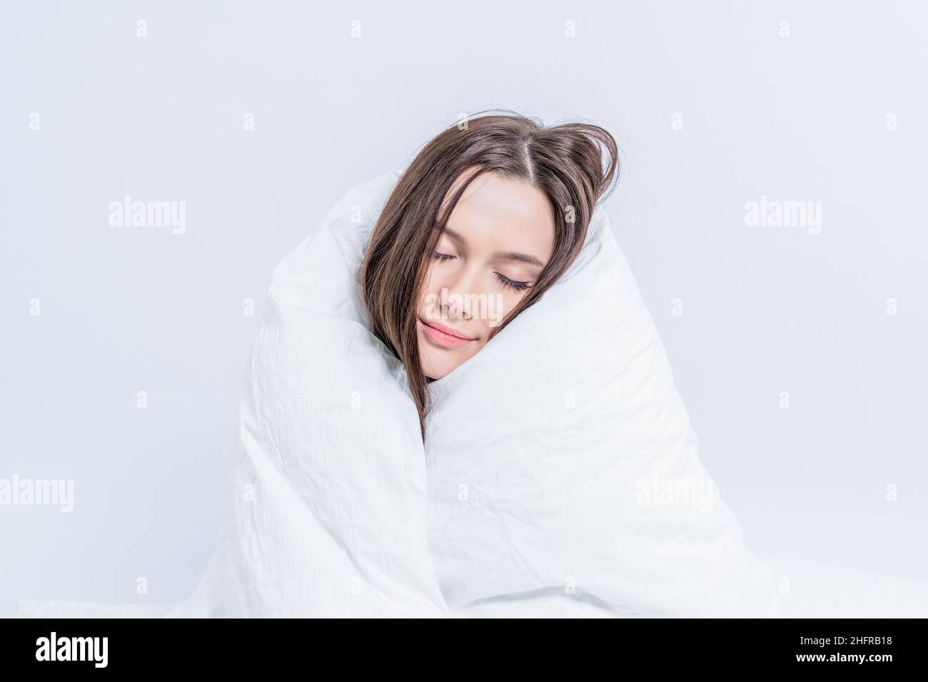 A pretty brunette, wrapped in a white blanket, sits on the bed. Keep warm with a warm blanket. Laziness. Stock Photo