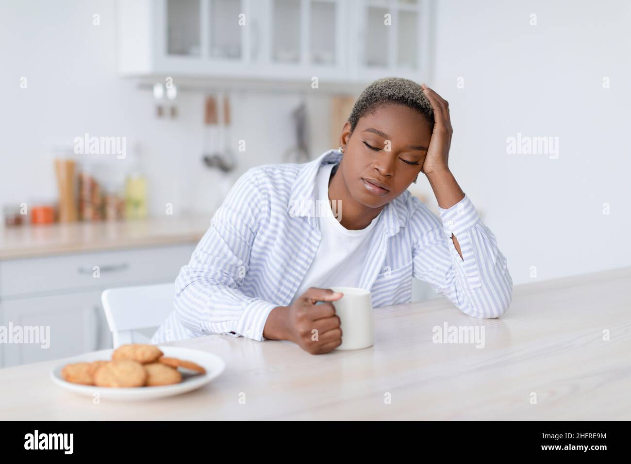 Tired sleepy young attractive black woman with cup of drink sits at table with cookies in scandinavian kitchen interior Stock Photo