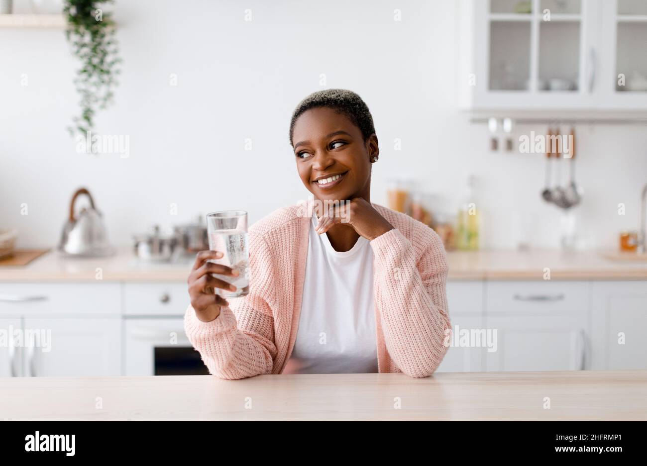 Smiling glad young attractive black lady holds glass of clean water, pensive and look at free space Stock Photo