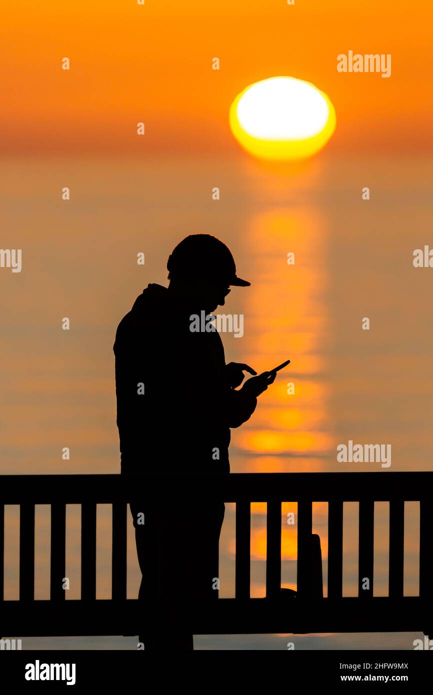 Aberystwyth, Ceredigion, Wales, UK. 17th January 2022  UK Weather: A silhouetted person looking at their phone, as the sun sets over the west coast of Aberystwyth. Wales. © Ian Jones/Alamy Live News Stock Photo