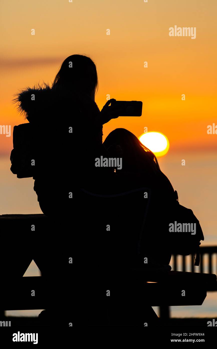 Aberystwyth, Ceredigion, Wales, UK. 17th January 2022  UK Weather: Silhouetted people sitting on a bench watching the sun setting over the west coast of Aberystwyth. Wales. © Ian Jones/Alamy Live News Stock Photo