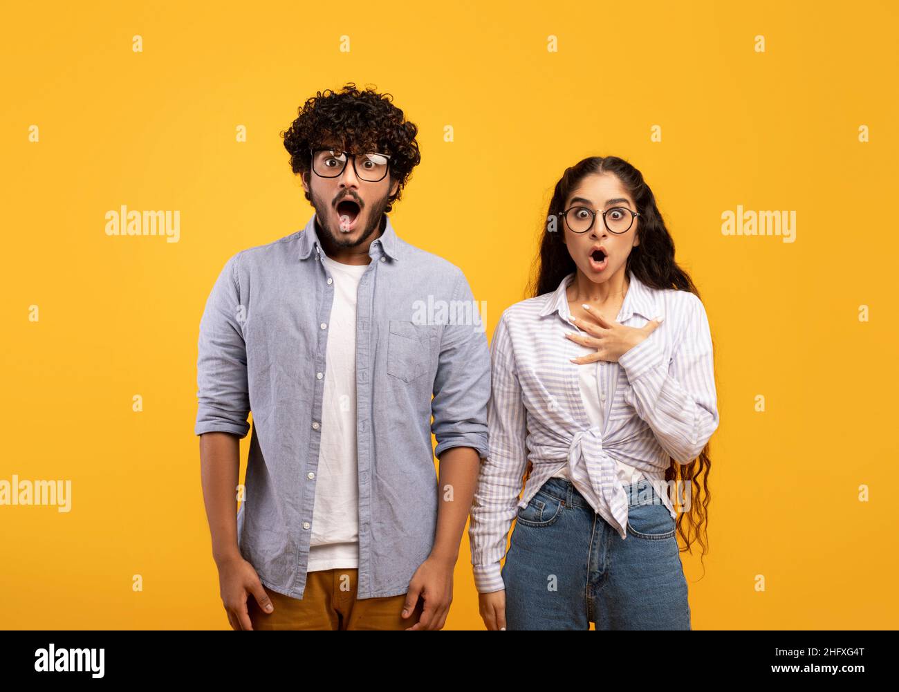 Shocked indian couple opening mouths in shock, woman putting hand on heart, standing over yellow studio background Stock Photo