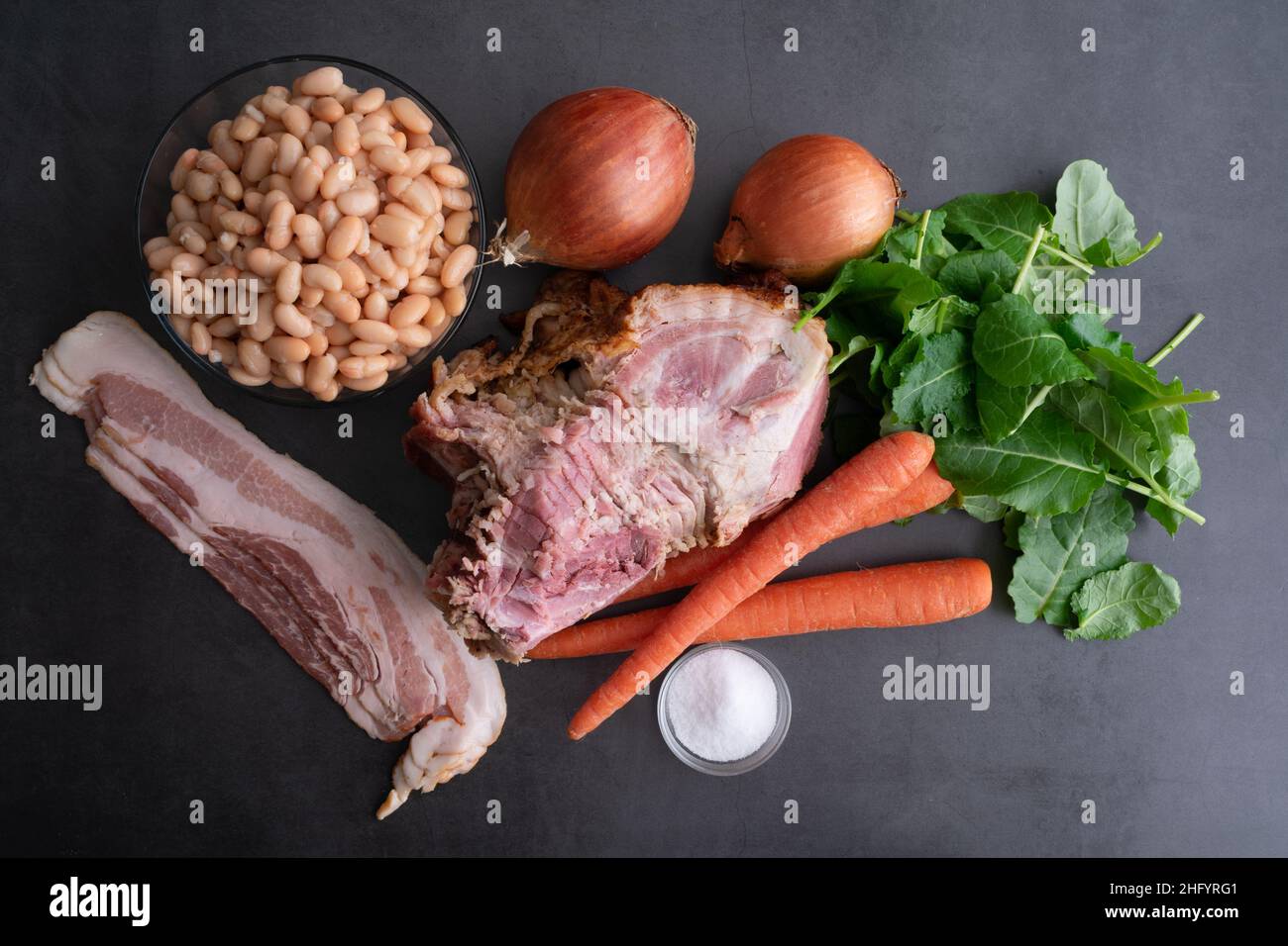 Ham Bone Soup with White Beans and Kale  Ingredients: Overhead view of hambone, bacon, and vegetables on a dark background Stock Photo
