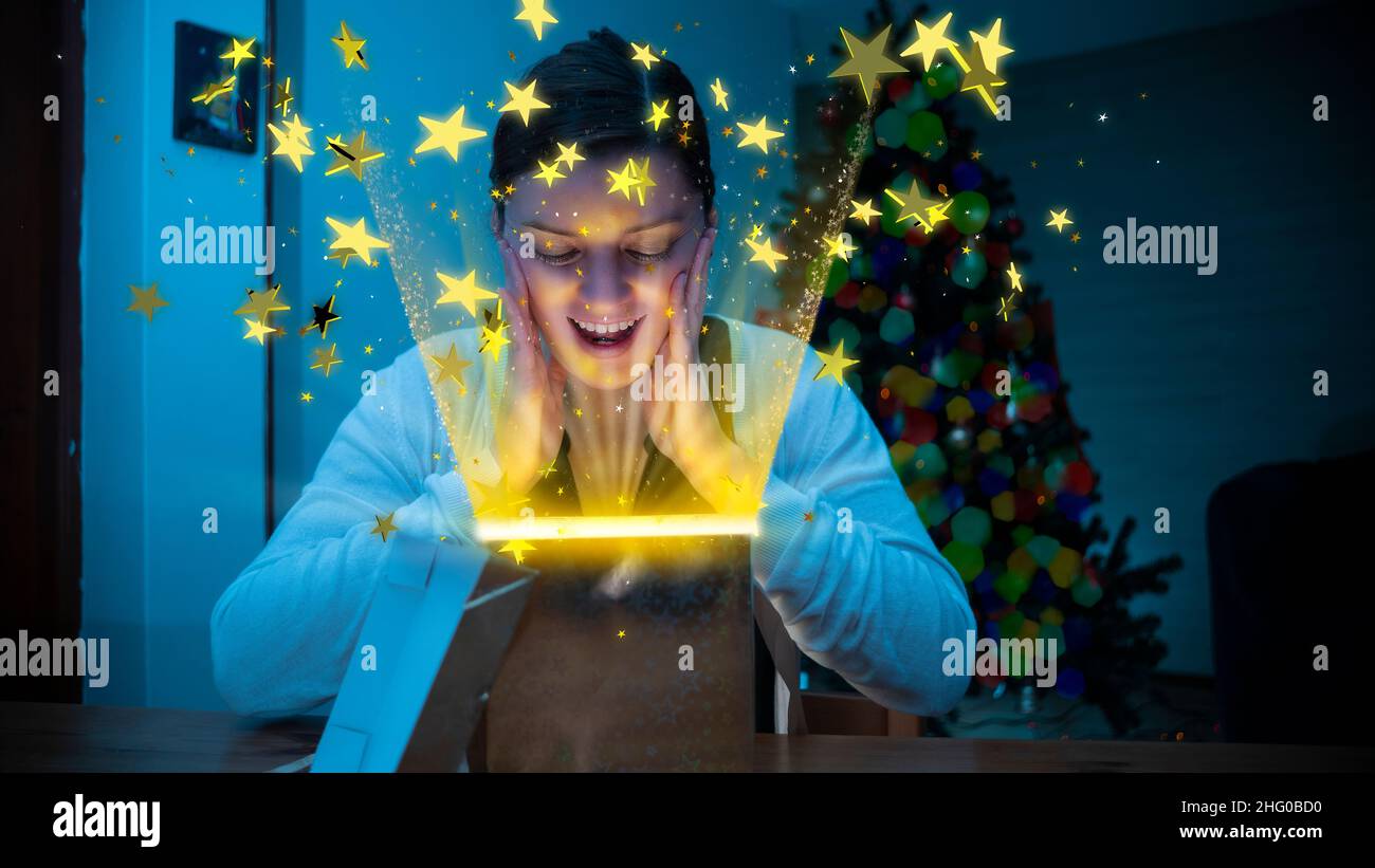 Beautiful Hispanic woman opening a gift box on a wooden table where a shower of stars comes out with yellow lighting from inside the box against a def Stock Photo