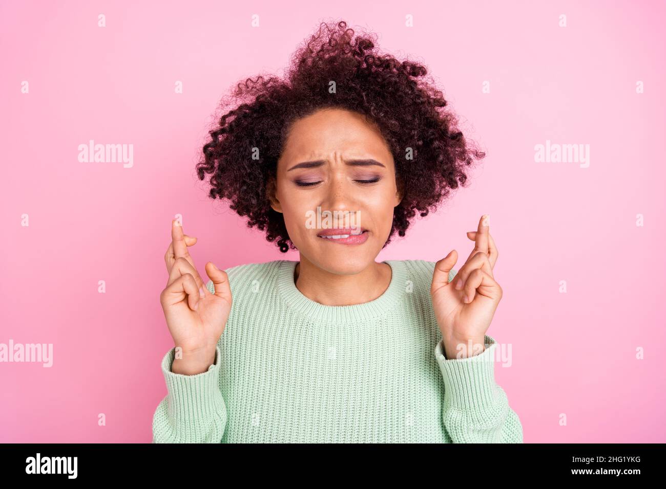 Portrait of pretty nervous dark skin lady closed eyes biting lip crossed fingers isolated on pink color background Stock Photo